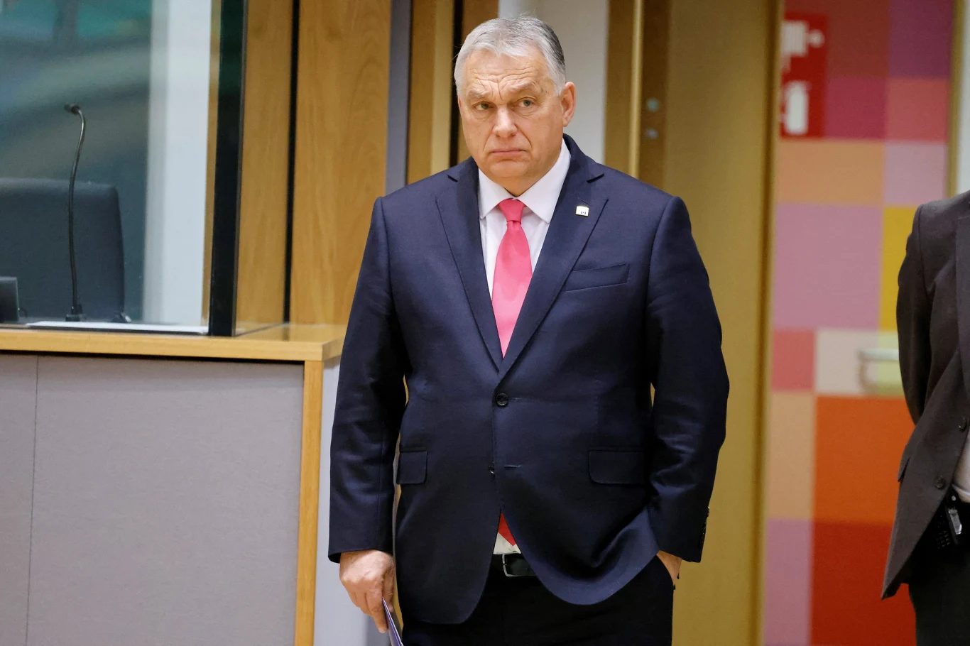 Viktor Orban od początku pełnoskalowej wojny w Ukrainie sprzeciwia się pomocy Kijowowi Mężczyzna w średnim wieku ubrany w ciemnogranatowy garnitur, biały koszula i różowy krawat stoi w nowoczesnym wnętrzu biurowym, obok drewnianych paneli i kolorowej ściany.