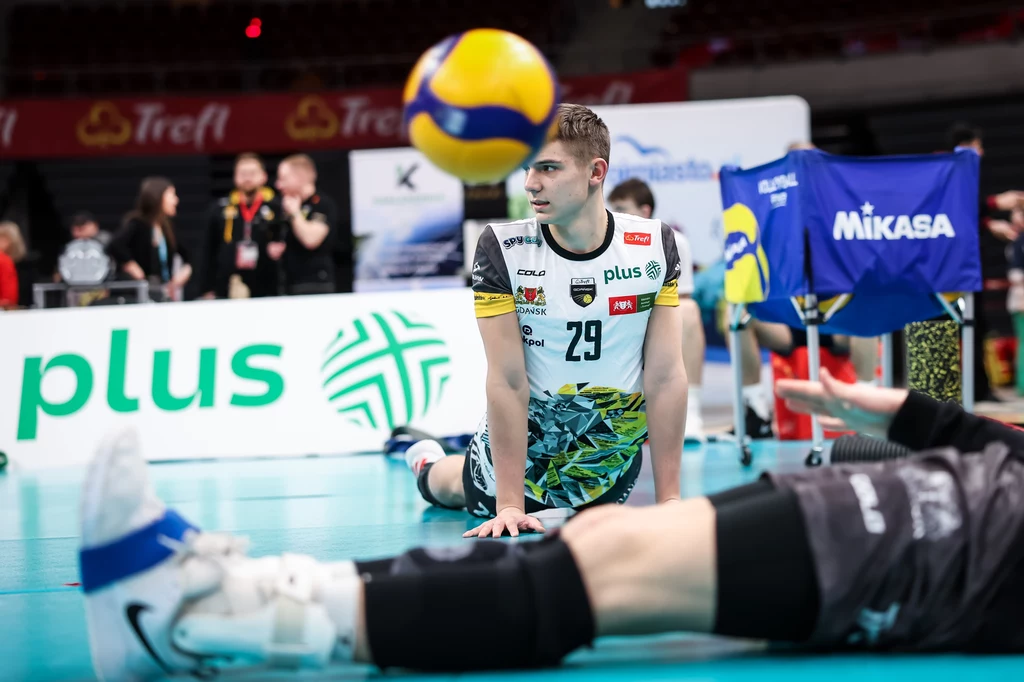 Michał Grzyb, jeden z filarów reprezentacji U-21 Siatkarz w sportowym stroju siedzący na parkiecie podczas rozgrzewki, w tle sprzęt treningowy, widoczne plansze sponsorów oraz unosząca się piłka siatkowa.