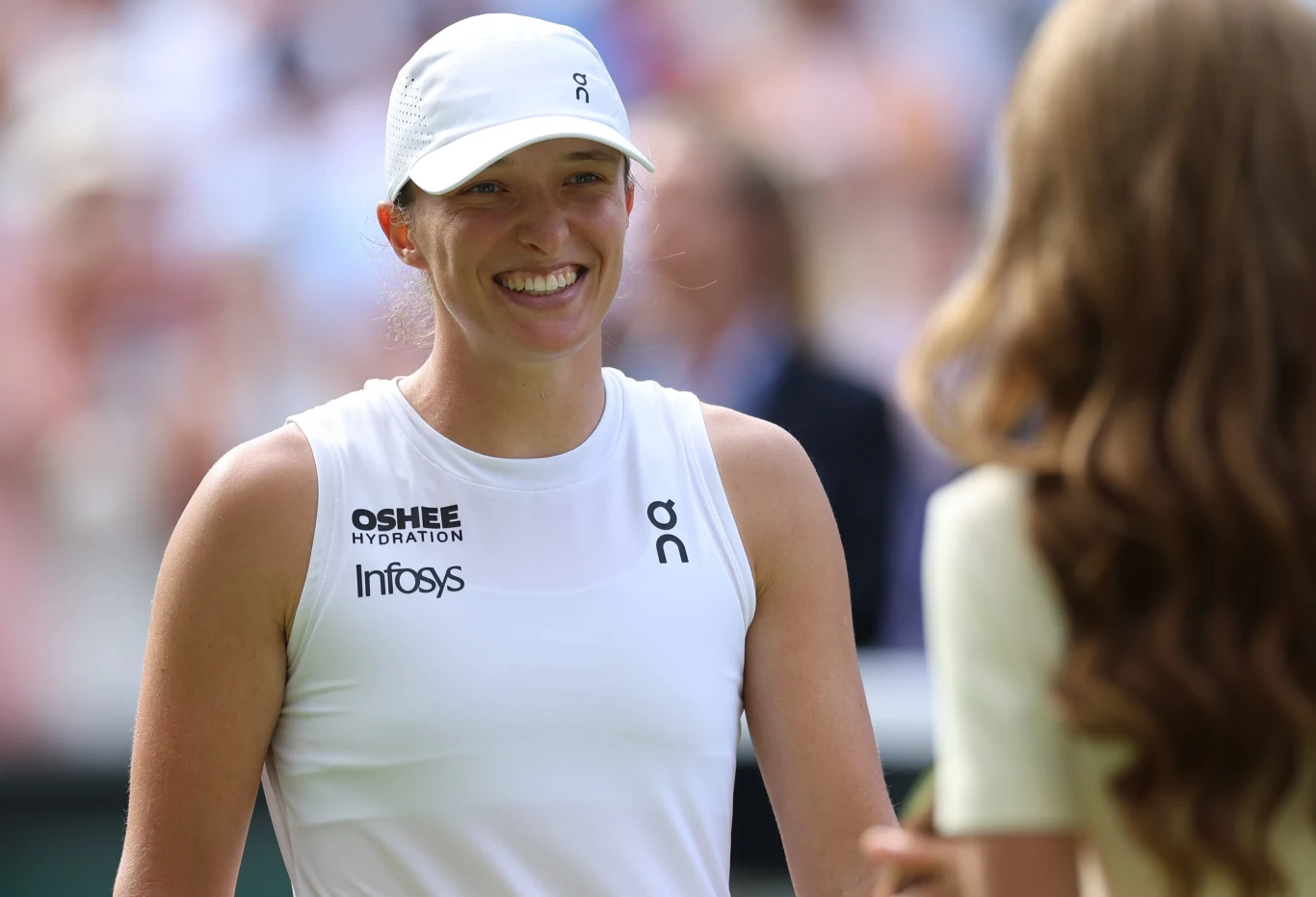 Tenisistka ubrana w biały strój sportowy i czapkę z daszkiem uśmiecha się, stojąc na korcie podczas rozmowy z inną osobą na pierwszym planie.