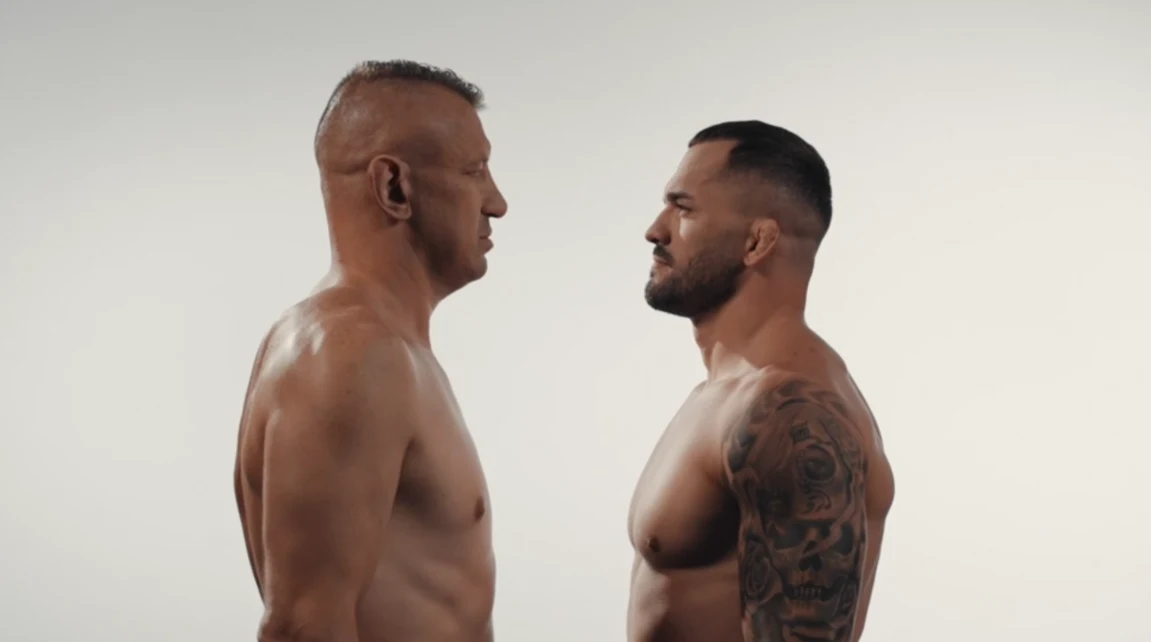 Dwóch umięśnionych mężczyzn stoi naprzeciwko siebie twarzą w twarz, bez koszulek, w postawie sugerującej gotowość do pojedynku, na jasnym, neutralnym tle.