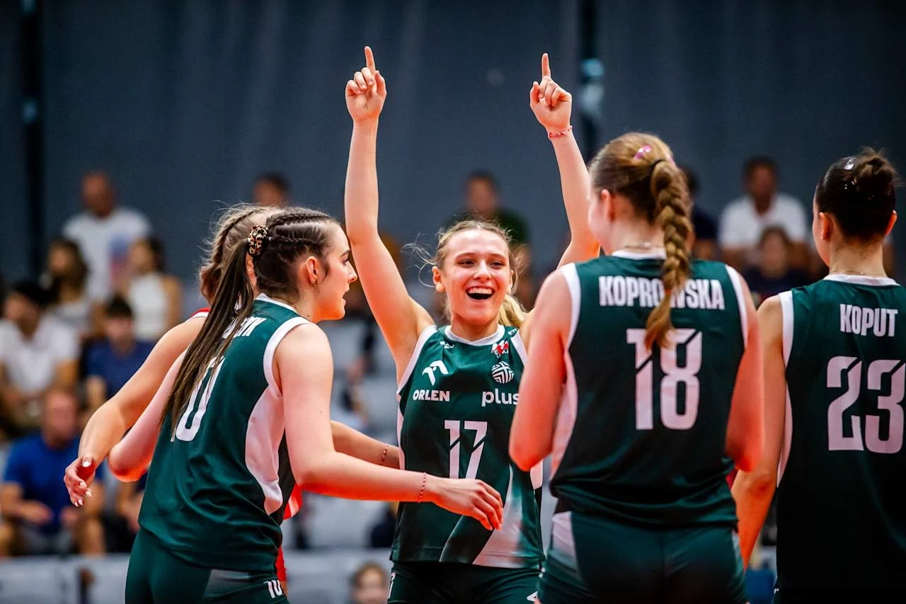 Grupa siatkarek w zielonych strojach sportowych świętuje punkt podczas meczu, jedna z zawodniczek unosi obie ręce do góry, a pozostałe wyrażają radość i entuzjazm, w tle widoczni są widzowie.