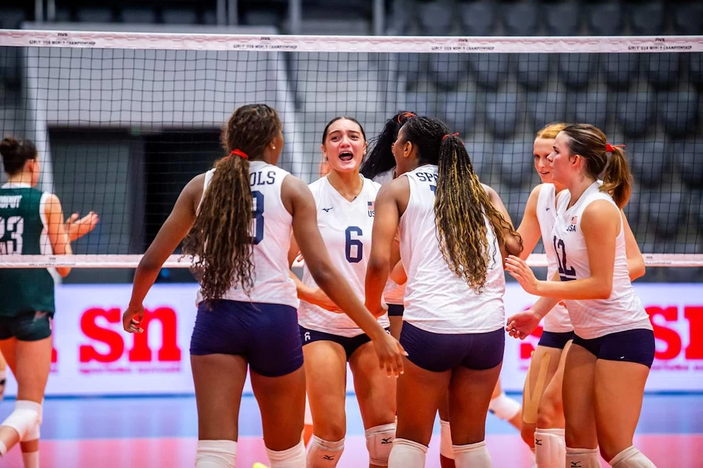 Siatkarki reprezentacji Polski U-19 Grupa siatkarek w białych koszulkach i granatowych spodenkach cieszy się po zdobyciu punktu na boisku, otaczając się w kółku i okazując radość. Tło stanowią puste trybuny i tablica świetlna.