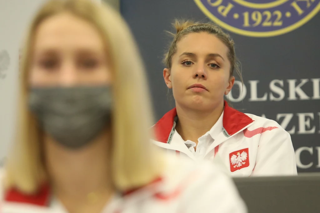 Alicja Tchórz Dwie kobiety w biało-czerwonych strojach reprezentacyjnych, jedna na pierwszym planie w maseczce ochronnej, druga na drugim planie z emblematem polskiego orła na ubraniu, w tle fragment logo związku sportowego.