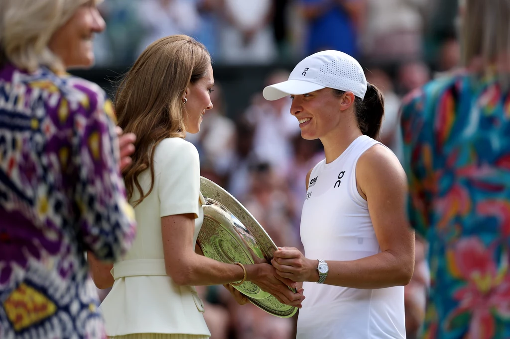 Iga Świątek i księżna Kate Zwyciężczyni turnieju tenisowego uśmiecha się i trzyma w rękach trofeum podczas uroczystości wręczenia nagród, naprzeciwko kobiety w eleganckiej, jasnej sukience, w tle rozmyty tłum widzów.