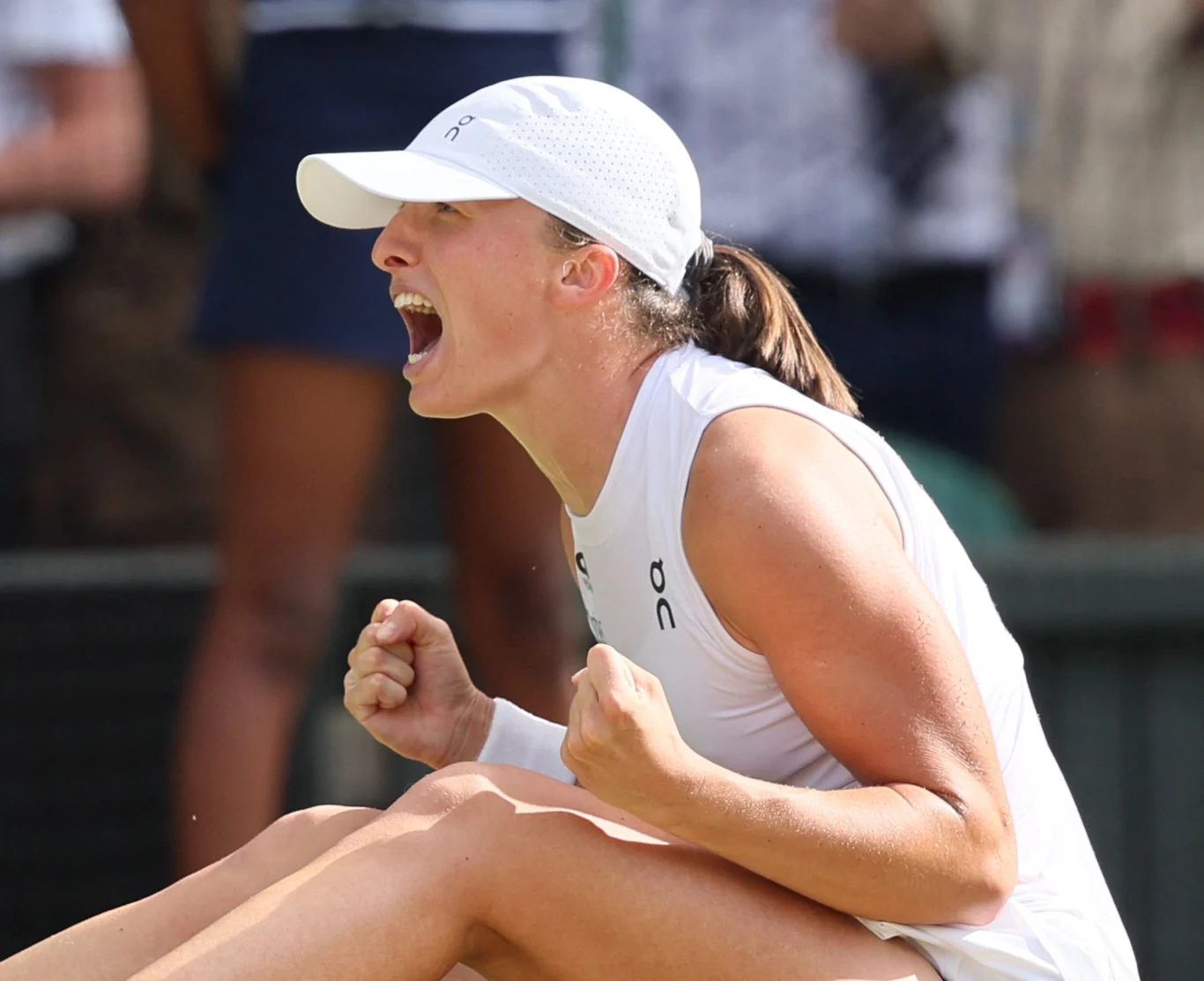 Tenisistka w białym stroju i czapce zaciska pięści, wyraża silne emocje i radość po udanym zagraniu, siedząc na korcie tenisowym.