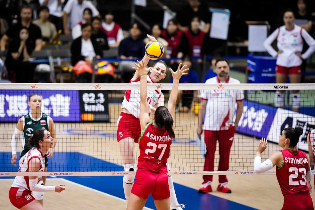 Liga Narodów siatkarek. Weronika Centka-Tietianiec w meczu Polska - Bułgaria Intensywna akcja podczas meczu siatkówki kobiet, zawodniczka w białym stroju atakuje piłkę nad siatką, naprzeciwko dwie zawodniczki w czerwonych strojach próbują zablokować atak, w tle widoczni trenerzy oraz obserwujący mecz zespół.