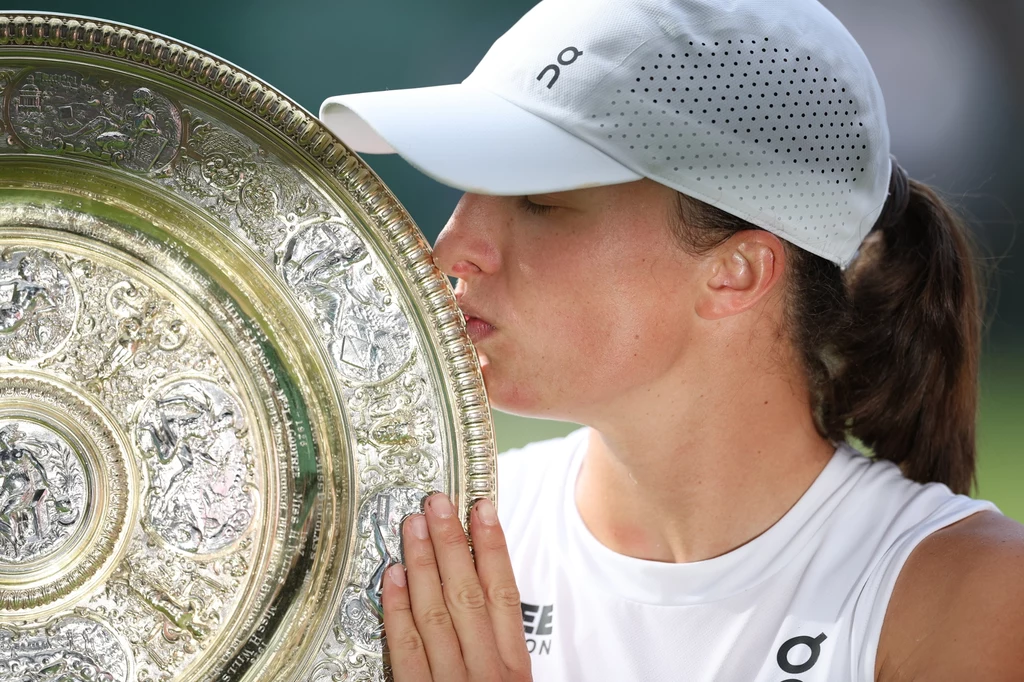 Iga Świątek z trofeum Wimbledonu Zawodniczka w białej czapce i stroju sportowym całuje herbowy złoty puchar o bogatym reliefie po zwycięstwie w prestiżowym turnieju tenisowym.
