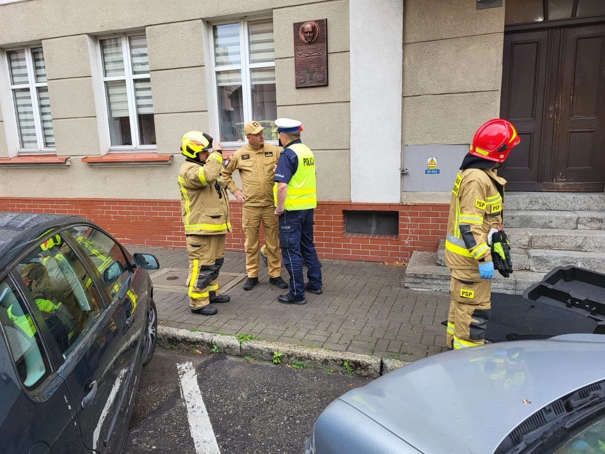 Potrącenie pieszych w Chełmnie Grupa strażaków i policjant rozmawiają na chodniku przy budynku z czerwoną ceglaną podmurówką, obok stoi zaparkowany samochód oraz samochód ze zdjętą maską, a jeden z ratowników trzyma wyposażenie ratownicze.
