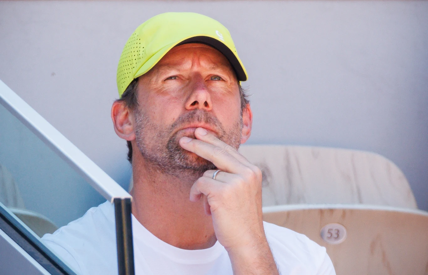 Mężczyzna w żółtej czapce z daszkiem, ubrany w biały T-shirt, siedzi na trybunach i zamyślony dotyka palcami brody.