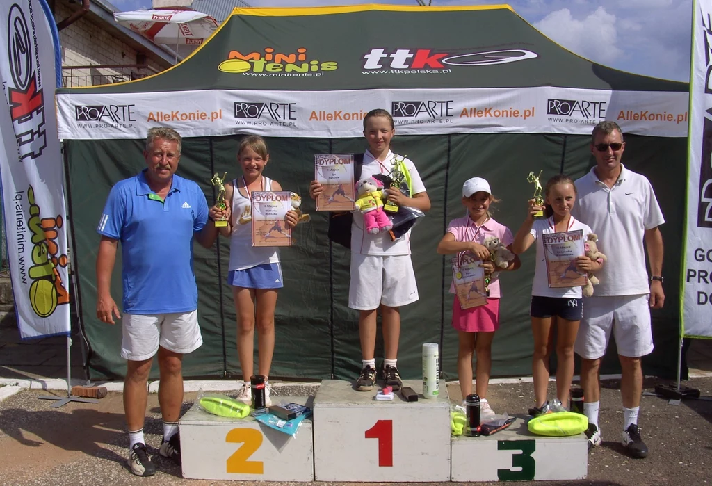 Grupa dzieci i dwóch dorosłych stoi na podium ze zdobytymi pucharami i dyplomami; w tle namiot z banerami sponsorskimi, dzieci ubrane w sportowe stroje tenisowe, na podium oznaczenia miejsc 1, 2, 3.