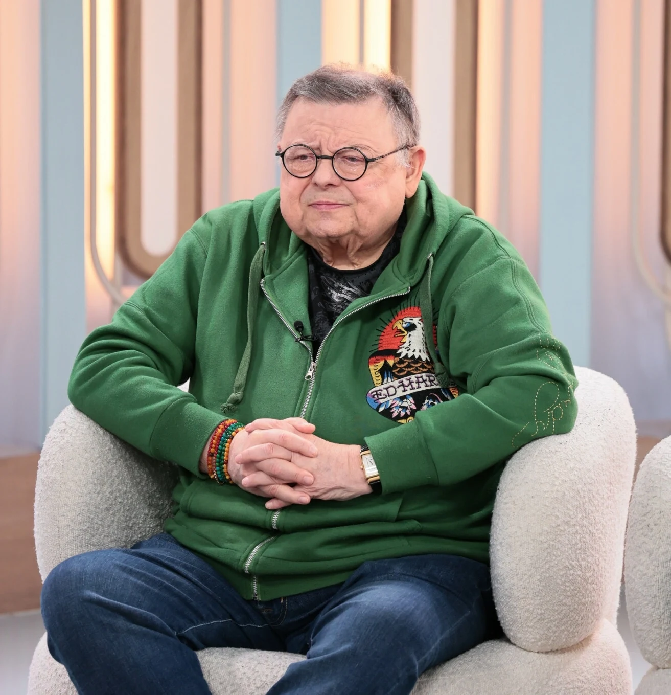 Starszy mężczyzna w okrągłych okularach, ubrany w zieloną bluzę z kolorowym wzorem i dżinsy, siedzi na jasnym fotelu w nowoczesnym, jasnym wnętrzu z geometrycznym tłem.