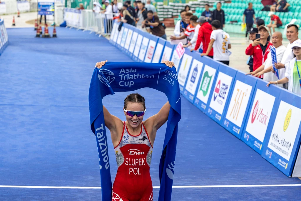 Roksana Słupek Triathlonistka w czerwonym stroju sportowym z napisem 'SLUPEK POL' przekracza linię mety, trzymając nad głową niebieską taśmę z logo Asia Triathlon Cup 2024, wokół dopingujący kibice oraz sędziowie i fotografowie.