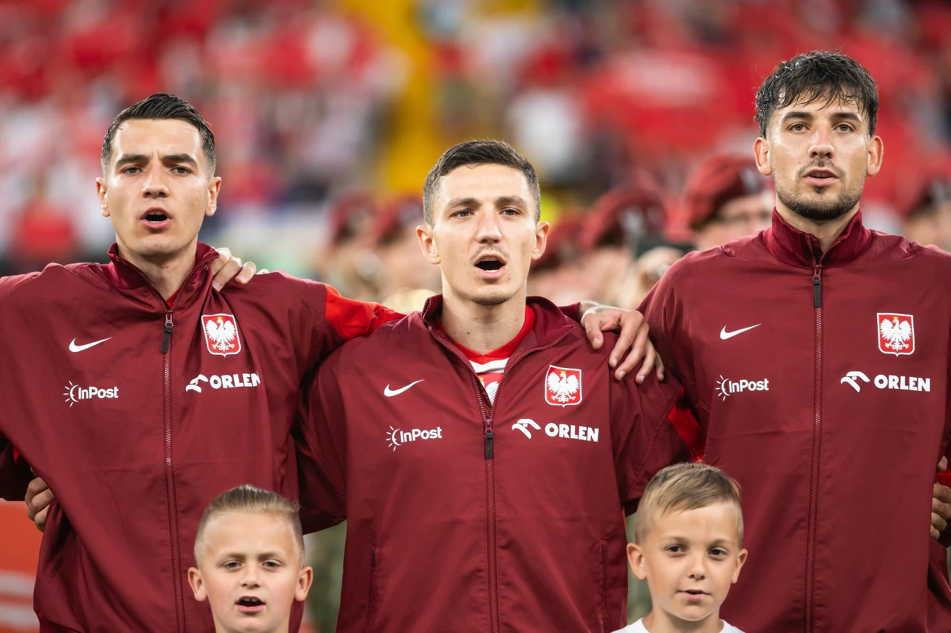 Jakub Kiwior (z lewej) Trzech piłkarzy w czerwonych kurtkach z godłem Polski stoi ramię w ramię podczas odśpiewywania hymnu narodowego, w tle widoczna rozmazana publiczność a przed nimi dwójka dzieci.