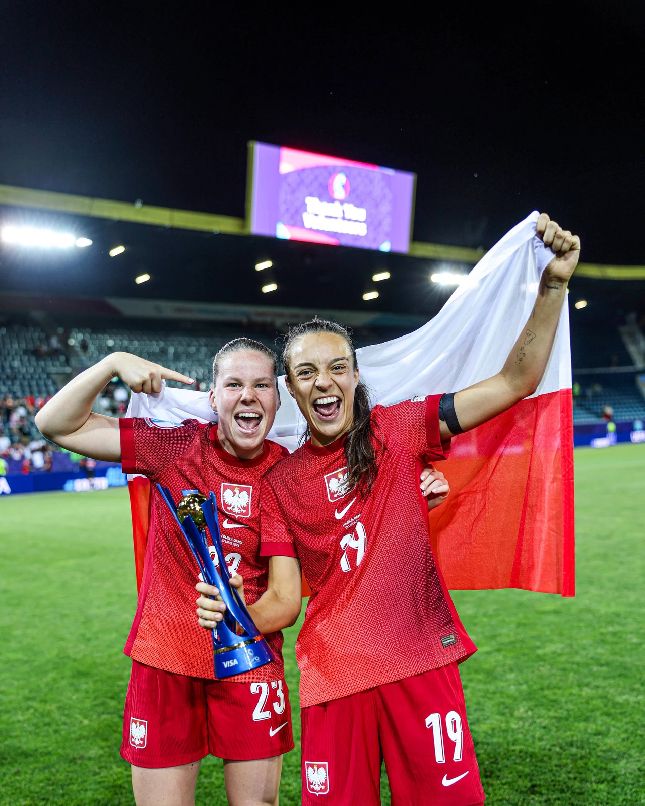 Dwie zawodniczki w czerwonych strojach z godłem Polski świętują na stadionie, trzymają polską flagę i puchar za zwycięstwo, uśmiechają się, w tle trybuny i tablica elektroniczna.