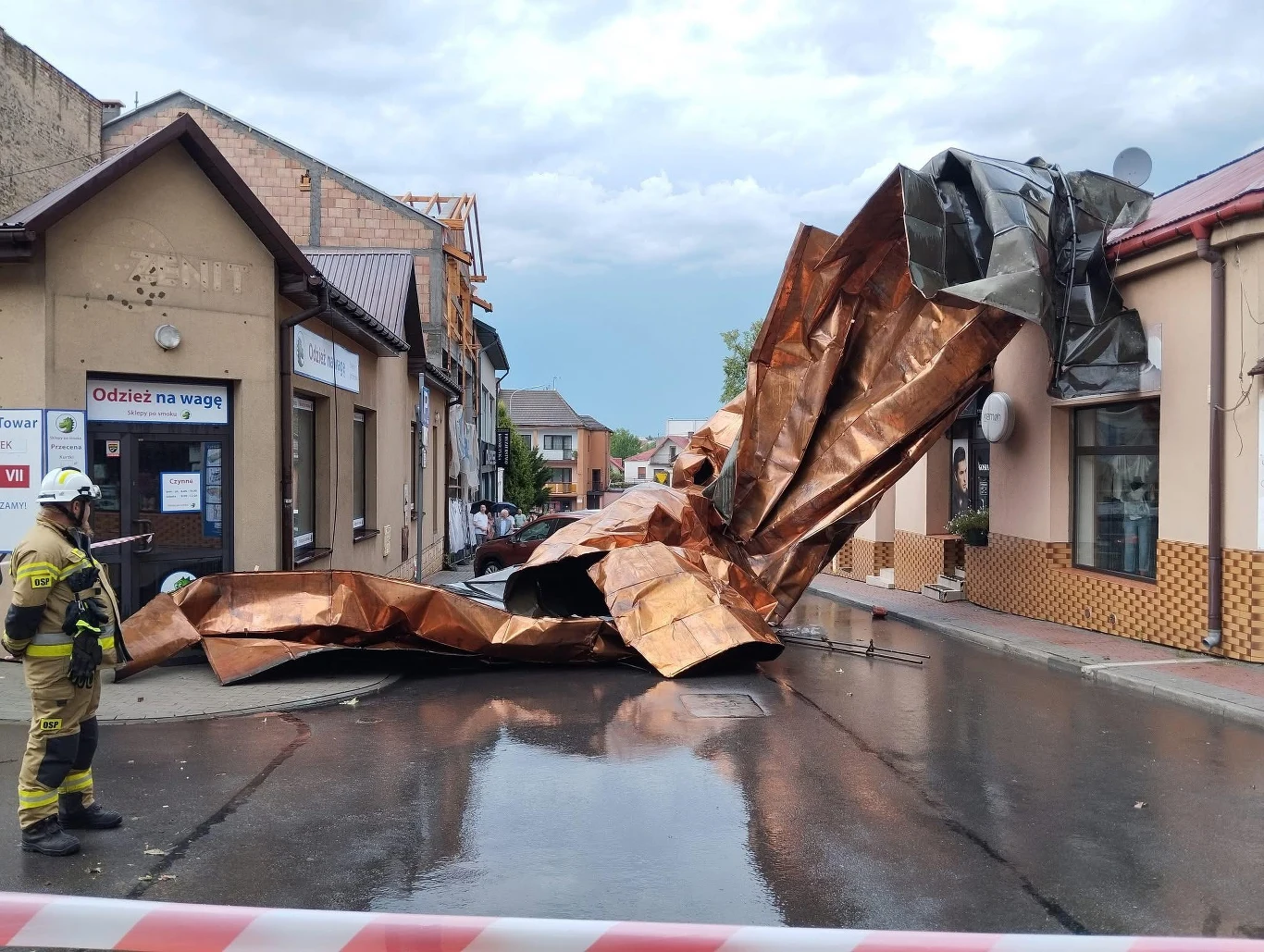 Zerwany dach leży na środku ulicy, otoczony przez budynki, jeden ze strażaków zabezpiecza miejsce zdarzenia, asfalt jest mokry od deszczu, a ulicę odgrodzono taśmą ostrzegawczą.