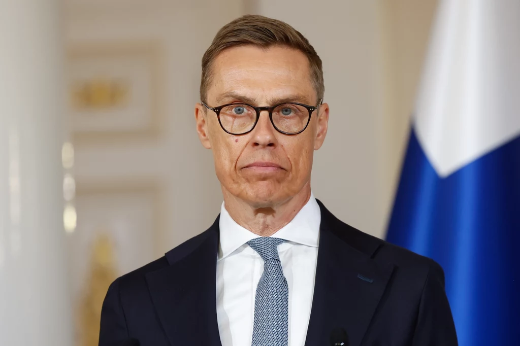 Alexander Stubb Mężczyzna w garniturze i okularach, z krótkimi włosami, stoi na tle jasnego wnętrza oraz fragmentu niebiesko-białej flagi.