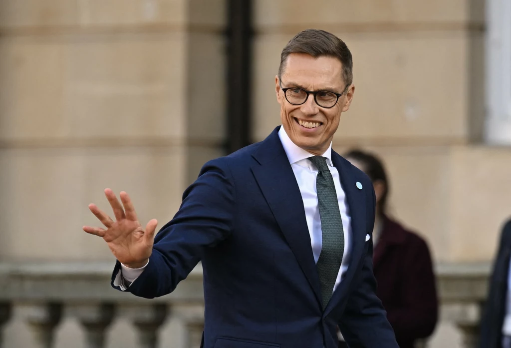 Alexander Stubb Elegancki mężczyzna w garniturze i okularach uśmiecha się i macha dłonią, stojąc na tle budynku o jasnej elewacji.