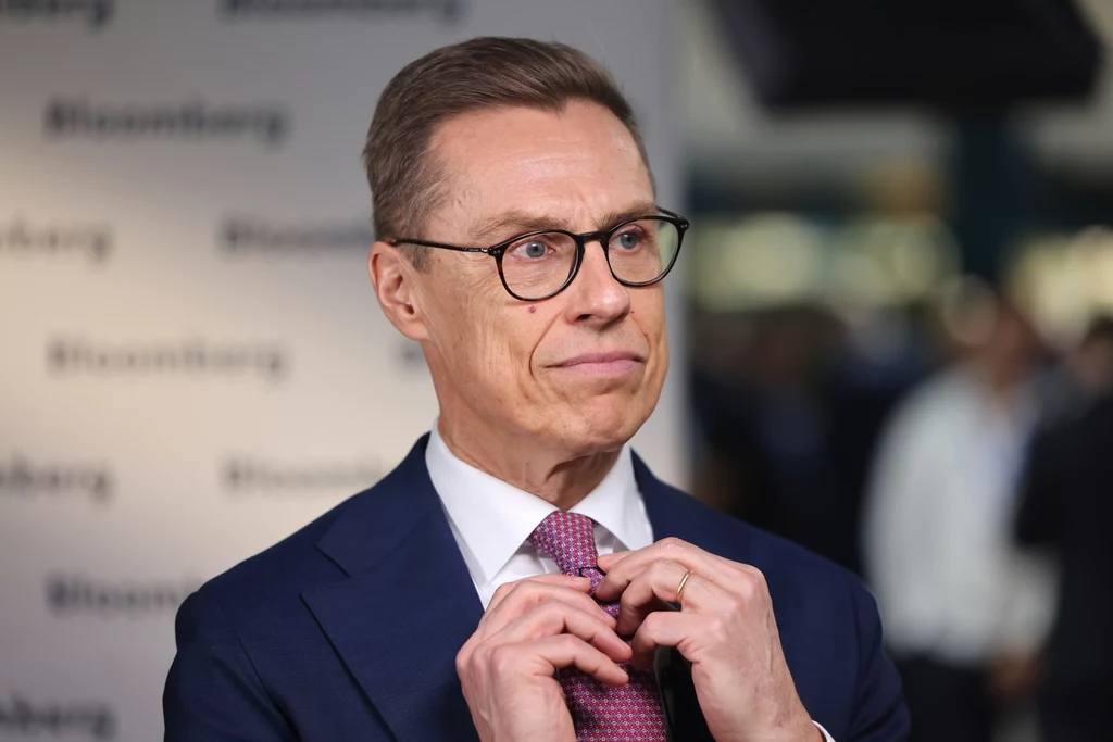 Alexander Stubb Mężczyzna w garniturze i okularach poprawia krawat na tle tablicy z powtarzającym się napisem Bloomberg, wyrażając skupienie i profesjonalizm.