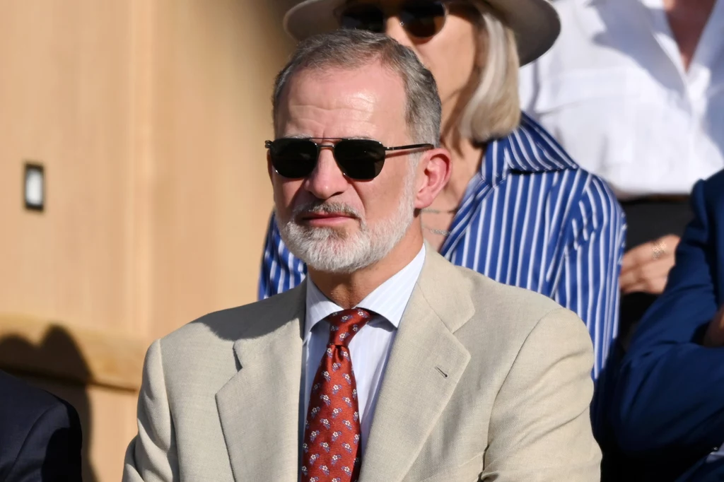 Król Filip VI na finale Wimbledonu Mężczyzna w jasnym garniturze, białej koszuli i czerwonym krawacie w granatowe wzory, z siwiejącą brodą i krótkimi włosami, w ciemnych okularach przeciwsłonecznych, siedzi na tle osób w eleganckich ubraniach.