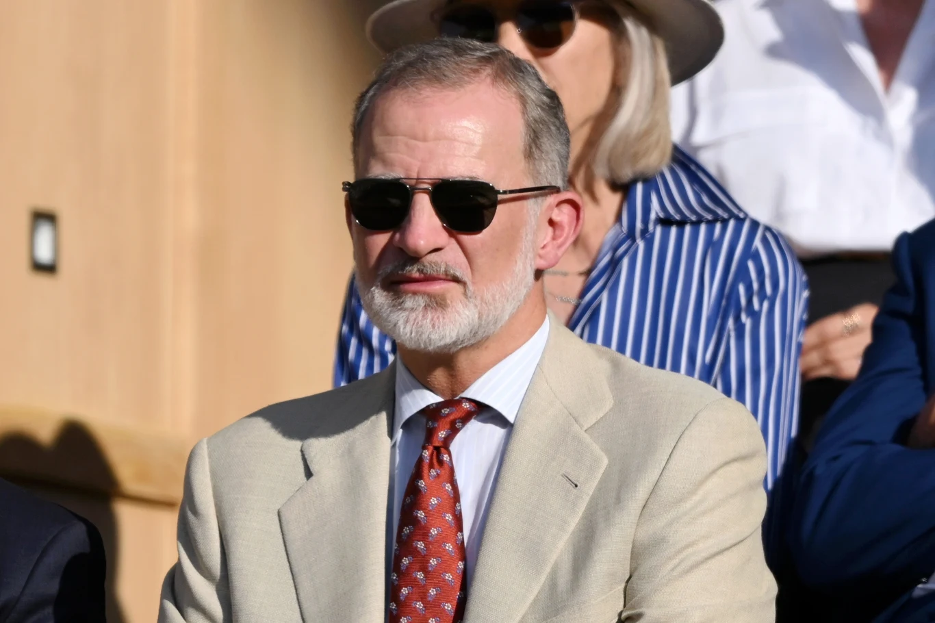 Mężczyzna w jasnym garniturze, białej koszuli i czerwonym krawacie w granatowe wzory, z siwiejącą brodą i krótkimi włosami, w ciemnych okularach przeciwsłonecznych, siedzi na tle osób w eleganckich ubraniach.