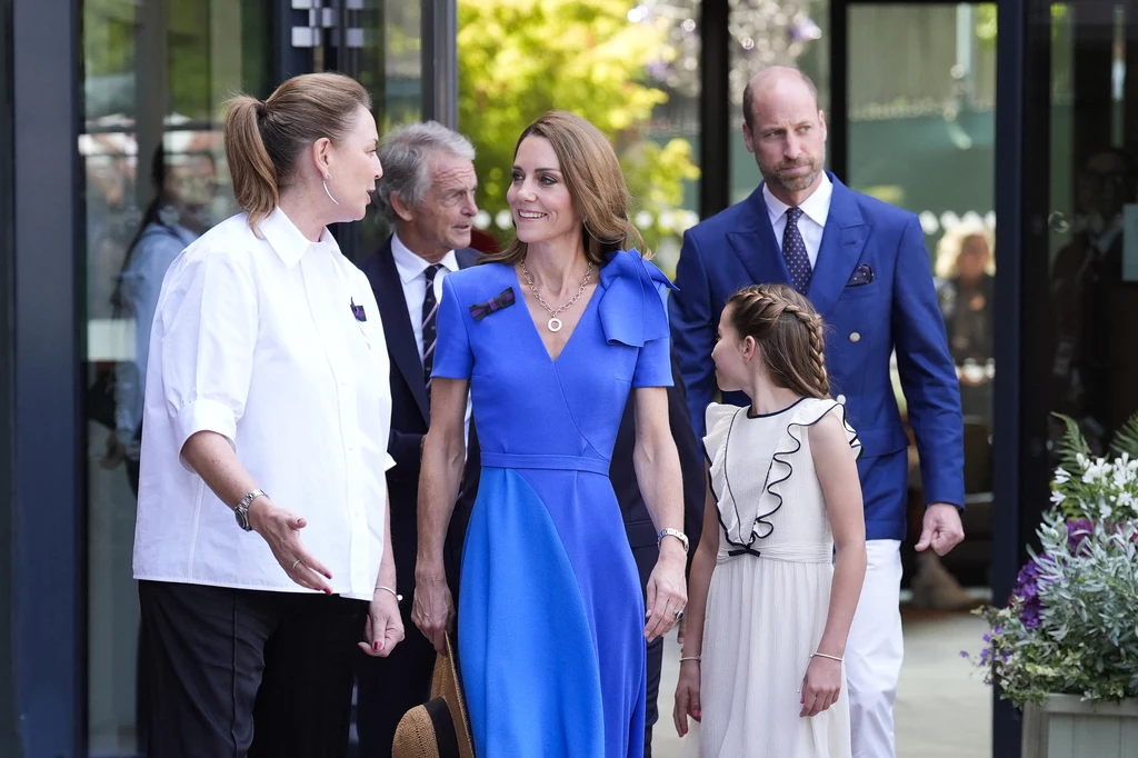 Księżna Kate z rodziną na finale Wimbledonu Grupa osób podczas oficjalnego wyjścia rozmawia na zewnątrz budynku, kobieta w eleganckiej niebieskiej sukni uśmiecha się do kobiety w białej bluzie kucharskiej, obok stoi dziewczynka w jasnej sukience i mężczyzna w granatowej marynarce