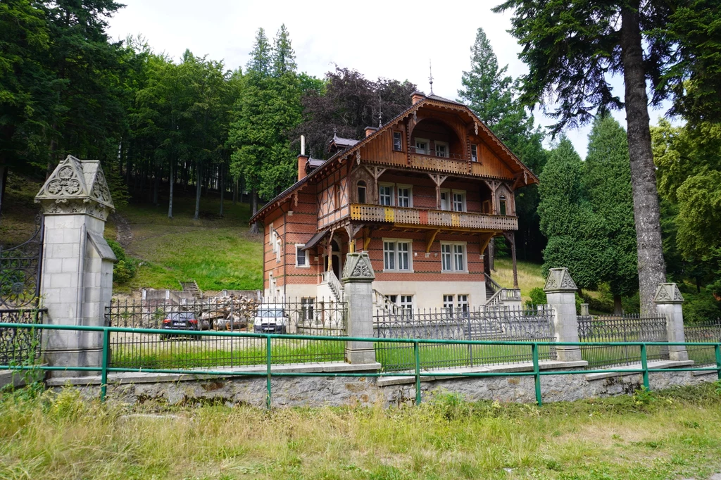 Leśny dworek w stylu tyrolskim w Bielawie to ciekawy zabytek architektoniczny. Od tego momentu szlak będzie prowadził przez spore wzniesienia Duży drewniany dom w stylu willowym, otoczony żeliwnym ogrodzeniem, zlokalizowany na tle lasu, z widocznymi samochodami zaparkowanymi obok budynku.