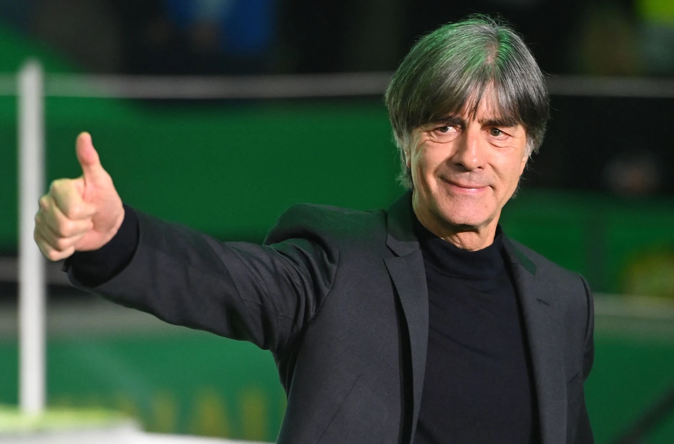 Mężczyzna w średnim wieku, ubrany w czarną marynarkę i koszulkę, pokazuje gest kciuka w górę, uśmiechając się lekko, w tle rozmyte światła oraz zielone elementy otoczenia.