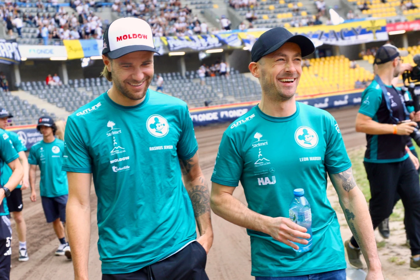 Rasmus Jensen i Leon Madsen Dwóch uśmiechniętych mężczyzn ubranych w turkusowe koszulki drużynowe idzie po stadionie żużlowym, wokół nich inni członkowie zespołu oraz kibice na trybunach; jeden z nich trzyma w ręku butelkę wody.