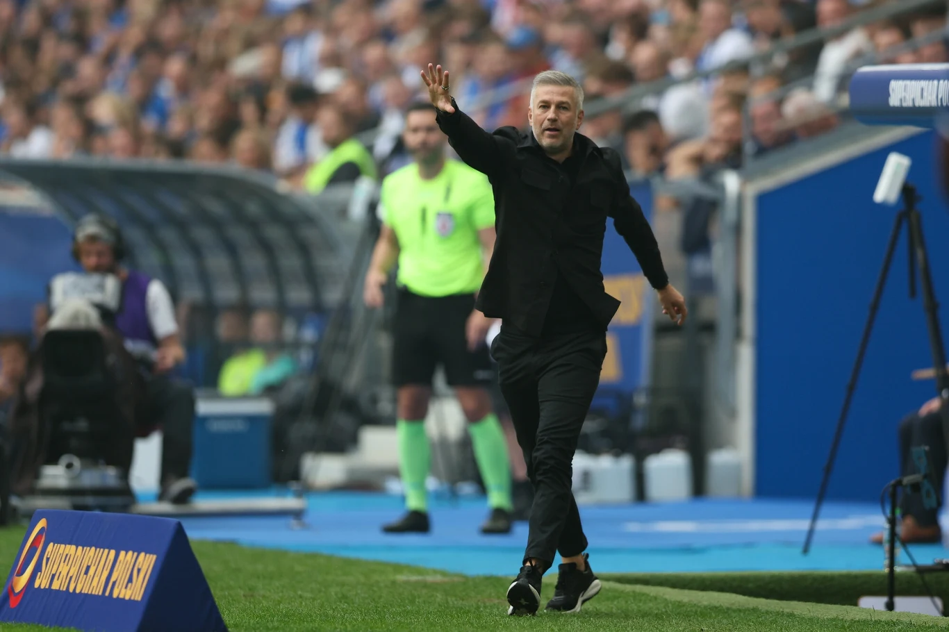 Trener Legii Warszawa Edward Iordanescu podczas meczu o Superpuchar Polski z Lechem Poznań Trener w ciemnym garniturze energicznie gestykuluje przy linii bocznej boiska podczas meczu piłkarskiego, w tle widać sędziego oraz zapełnione trybuny stadionu.