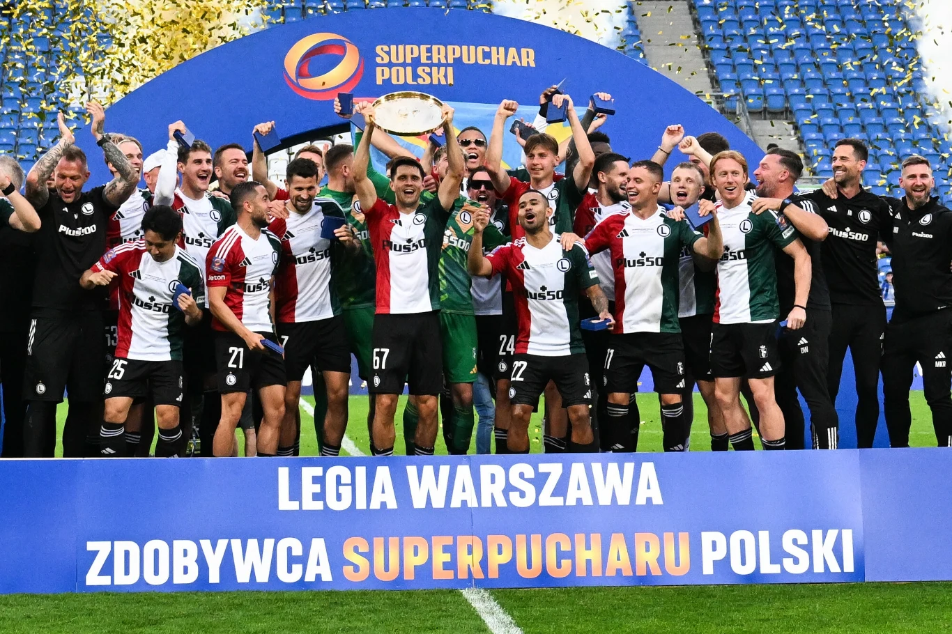 Piłkarze Legii Warszawa celebrują zwycięstwo w Superpucharze Polski na stadionie, trzymając trofeum i unosząc ręce w geście triumfu, otoczeni przez konfetti i baner z nazwą klubu oraz informacją o wygranej.