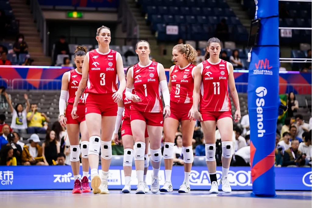 Siatkarki reprezentacji Polski Grupa siatkarek w czerwonych strojach, wyposażonych w ochraniacze na kolana, wchodzi na boisko podczas meczu halowego, wokół widoczni kibice na trybunach oraz sportowy sprzęt i elementy wyposażenia hali.