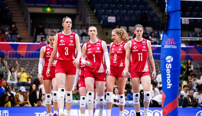Grupa siatkarek w czerwonych strojach, wyposażonych w ochraniacze na kolana, wchodzi na boisko podczas meczu halowego, wokół widoczni kibice na trybunach oraz sportowy sprzęt i elementy wyposażenia hali.