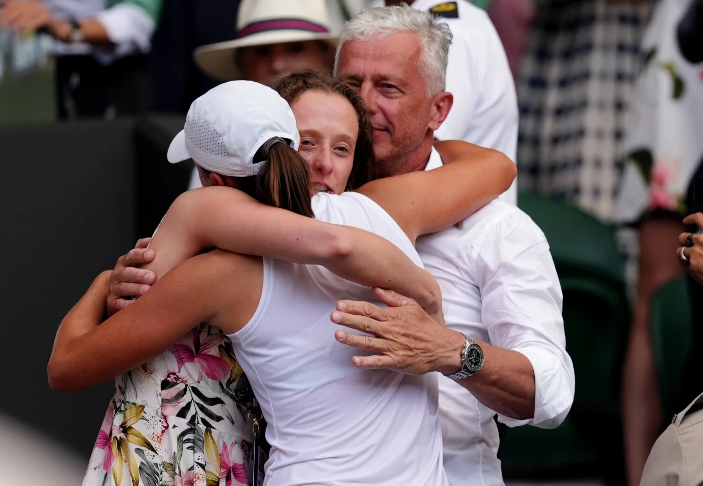 Trzy osoby obejmują się wzajemnie, wyrażając radość i wzruszenie, a w tle widać innych ludzi, którzy obserwują sytuację.
