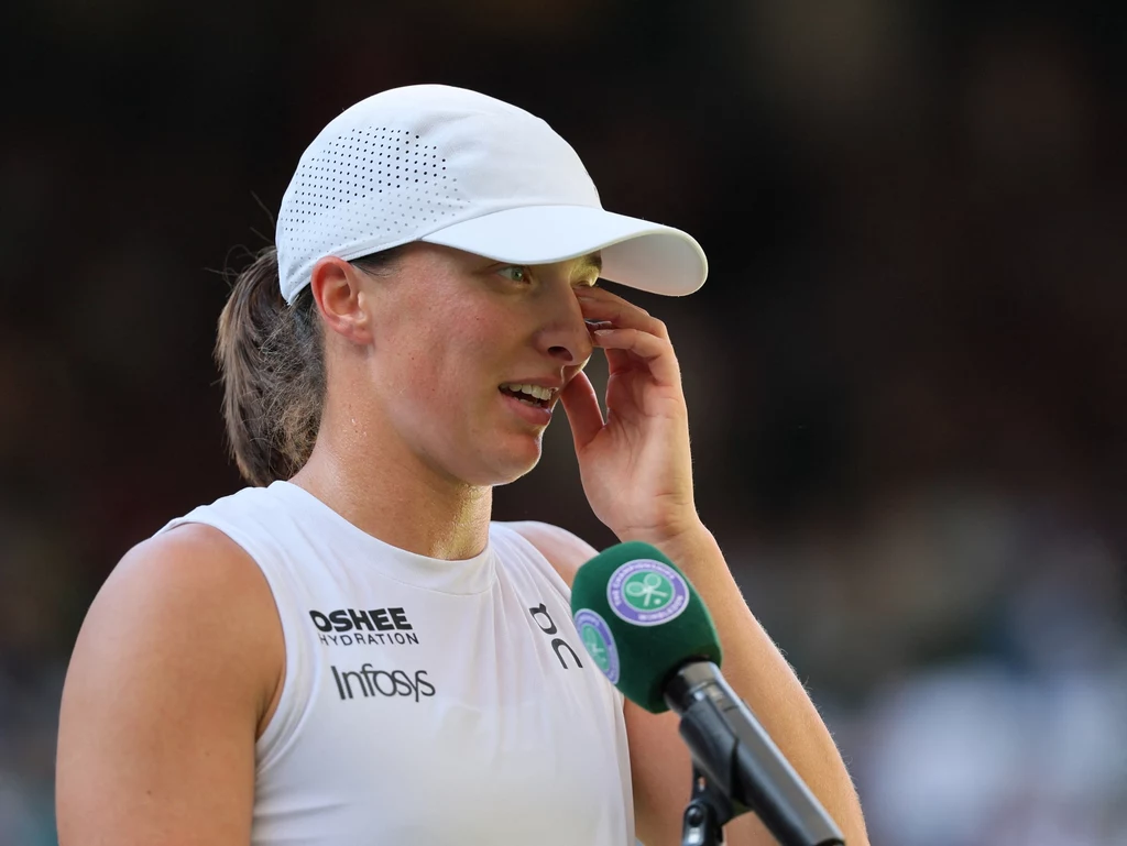 Iga Świątek z trudem powstrzymawa łzy po triumfie na Wimbledonie Kobieta w białej czapce z daszkiem i sportowej koszulce mówi do mikrofonu po meczu tenisowym, trzymając rękę przy twarzy; w tle rozmyte ciemne tło stadionu.