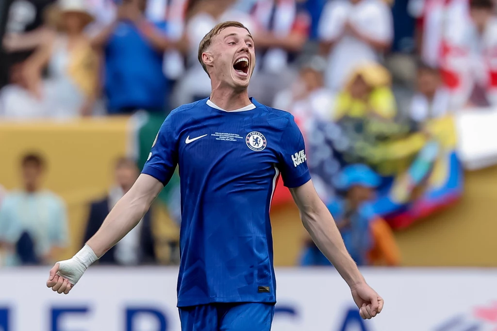 Cole Palmer Piłkarz w niebieskiej koszulce z herbem Chelsea świętuje na boisku, mając uniesione ręce i szeroko otwarte usta; w tle widać rozmytą publiczność.