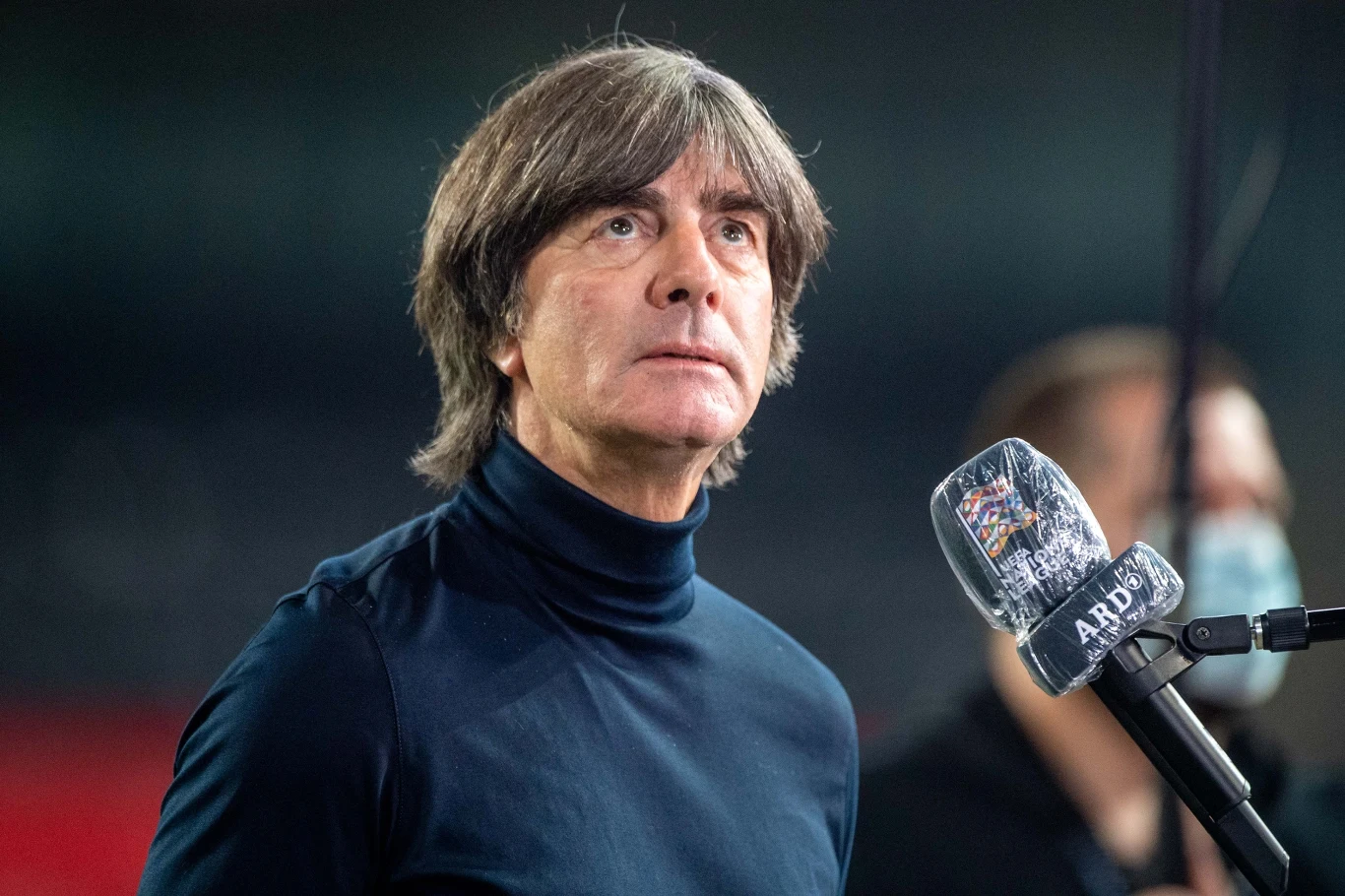 Mężczyzna o średniej długości ciemnych włosach w granatowym golfie stoi przed mikrofonem oznaczonym logo ARD, w tle nieostre sylwetki i neutralne oświetlenie stadionowe.