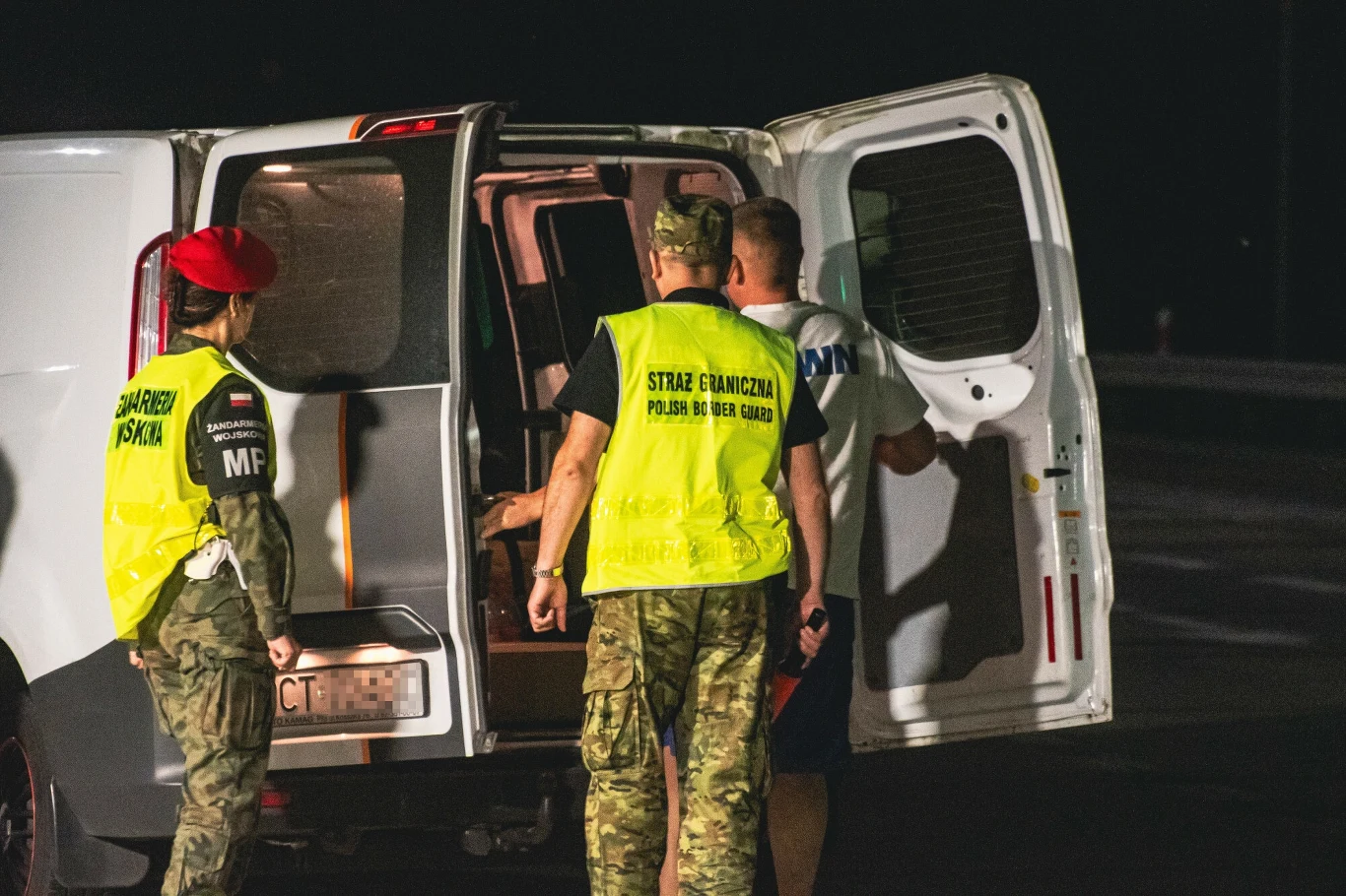 Funkcjonariusze Straży Granicznej w kamizelkach odblaskowych oraz mężczyzna w jasnej koszulce stoją przy otwartych drzwiach białego samochodu dostawczego w nocy.