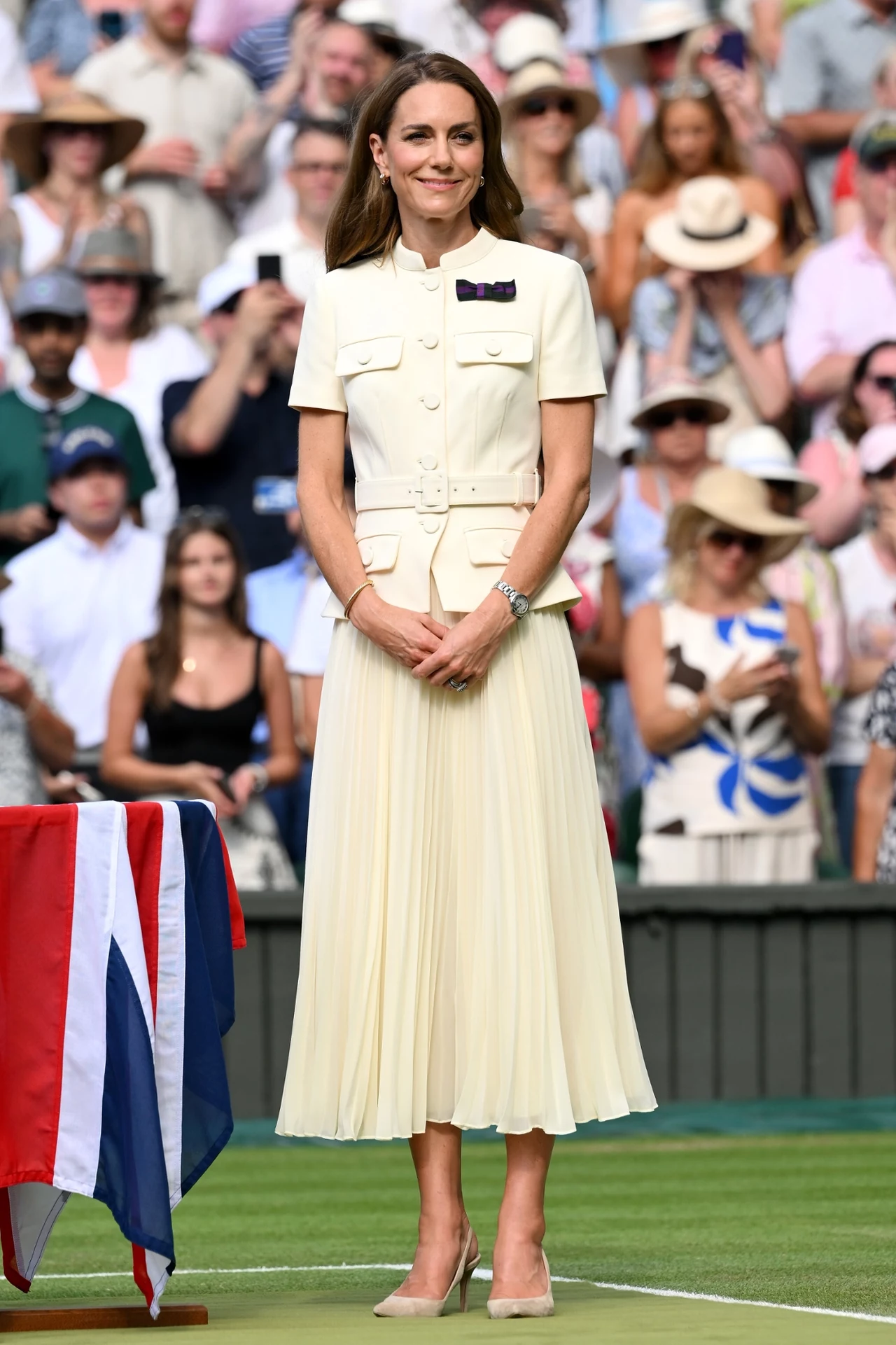 Kolor kreacji księżnej Kate to hit lata 2025 Elegancka kobieta w kremowym garniturze ze spódnicą o długości midi stoi na trawie w pobliżu stołu przykrytego brytyjską flagą; za nią tłum osób ubranych w letnie ubrania.