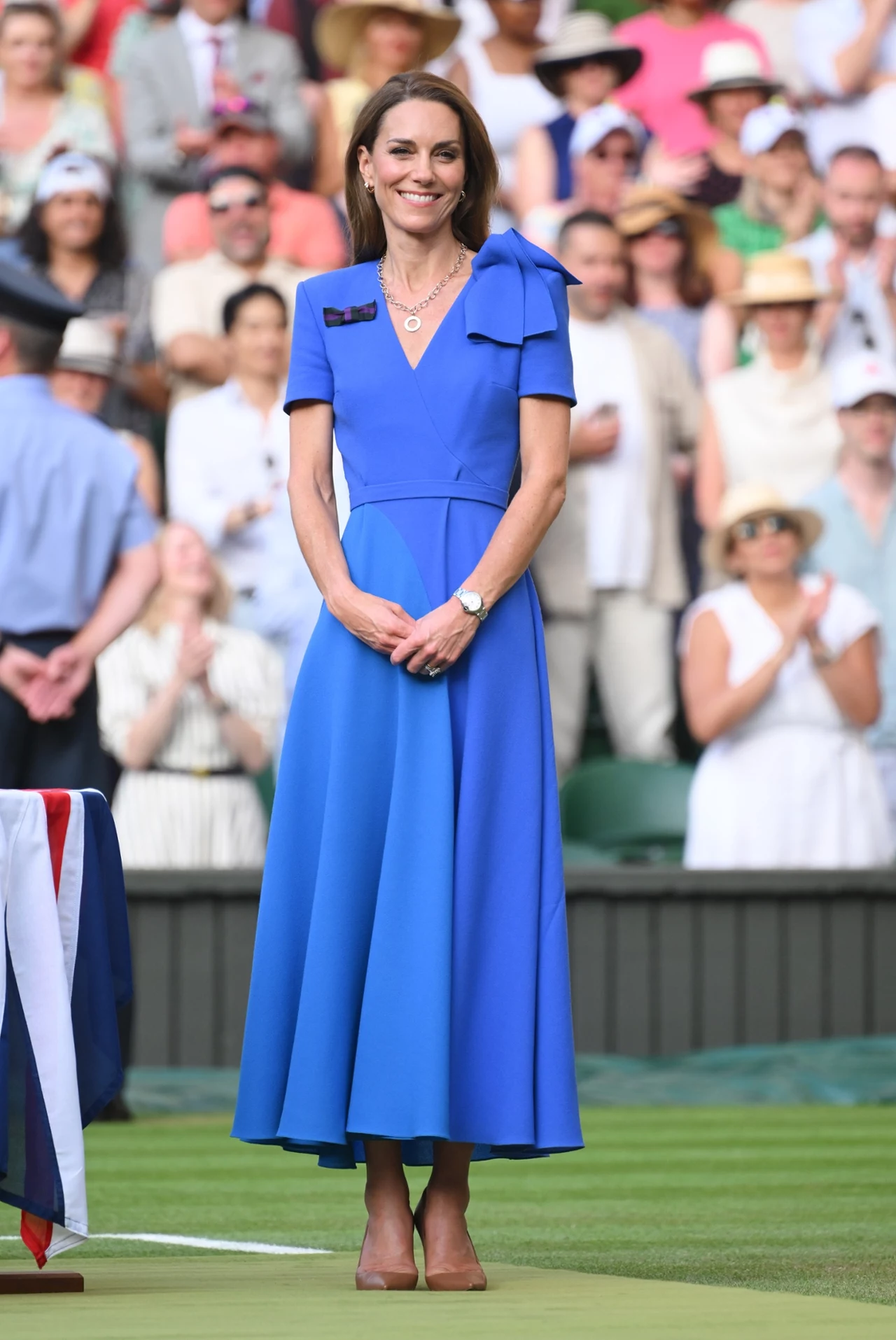 Druga kreacja Kate wyróżniała się krojem i zdecydowanym akcentem kolorystycznym Elegancko ubrana kobieta w niebieskiej sukience stoi na zielonym korcie tenisowym, w tle znajduje się tłum obserwujących osób, większość z nich nosi letnie ubrania i kapelusze.
