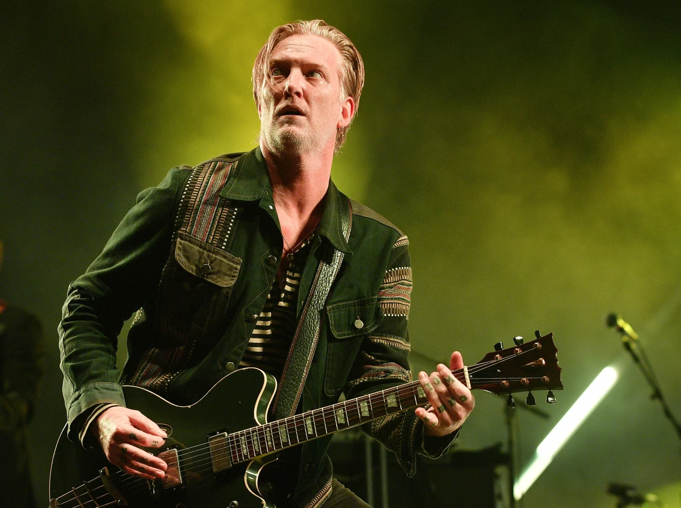 Josh Homme, frontman zespołu Queens of The Stone Agę Mężczyzna grający na gitarze elektrycznej podczas występu na scenie, w tle intensywne światła w odcieniach żółci i zieleni podkreślają koncertową atmosferę.