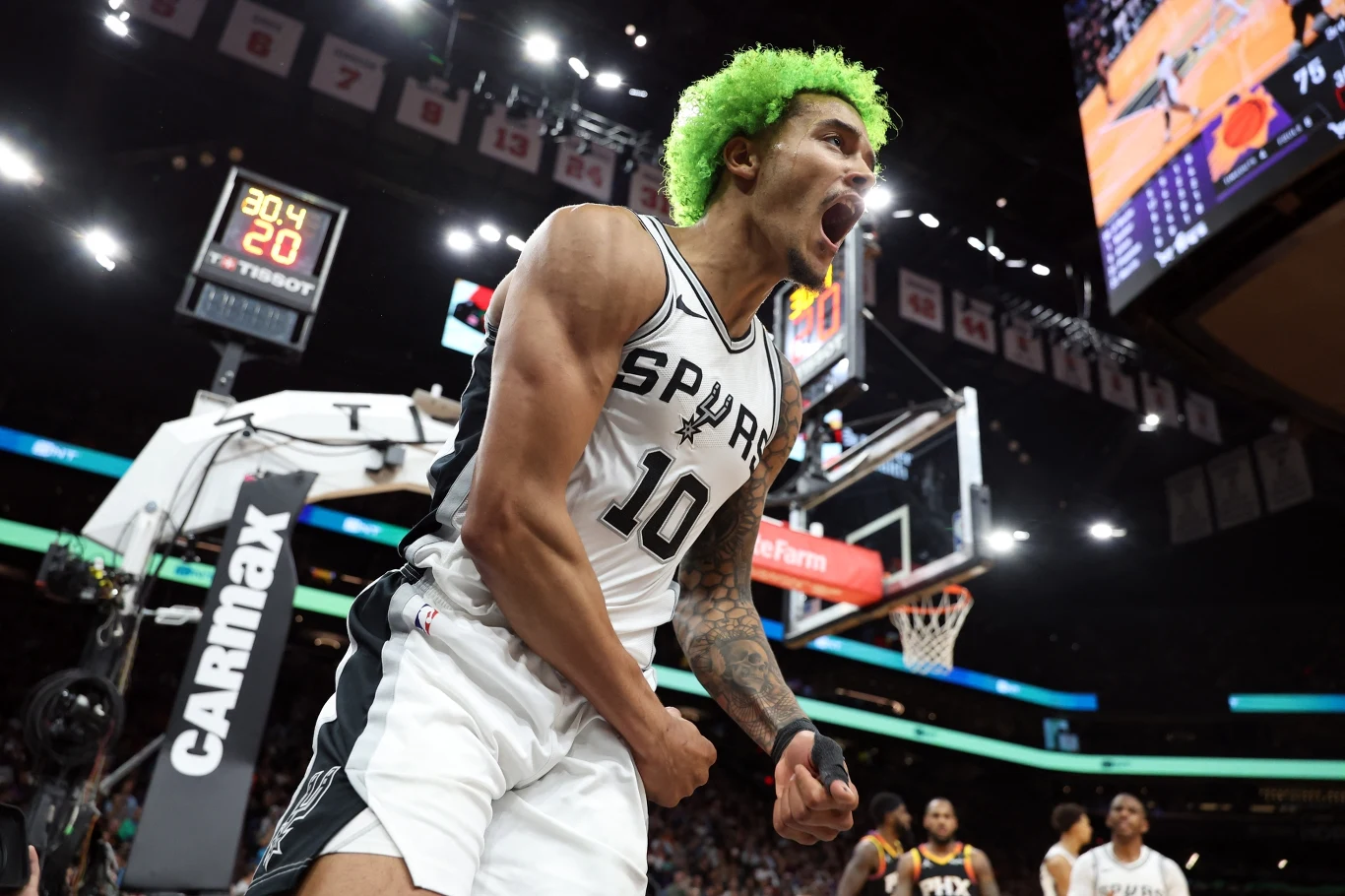 Koszykarz w stroju drużyny Spurs z zielonym afro energicznie świętuje na parkiecie podczas meczu, w tle widoczne są tablica wyników, kosz oraz inni zawodnicy na boisku.