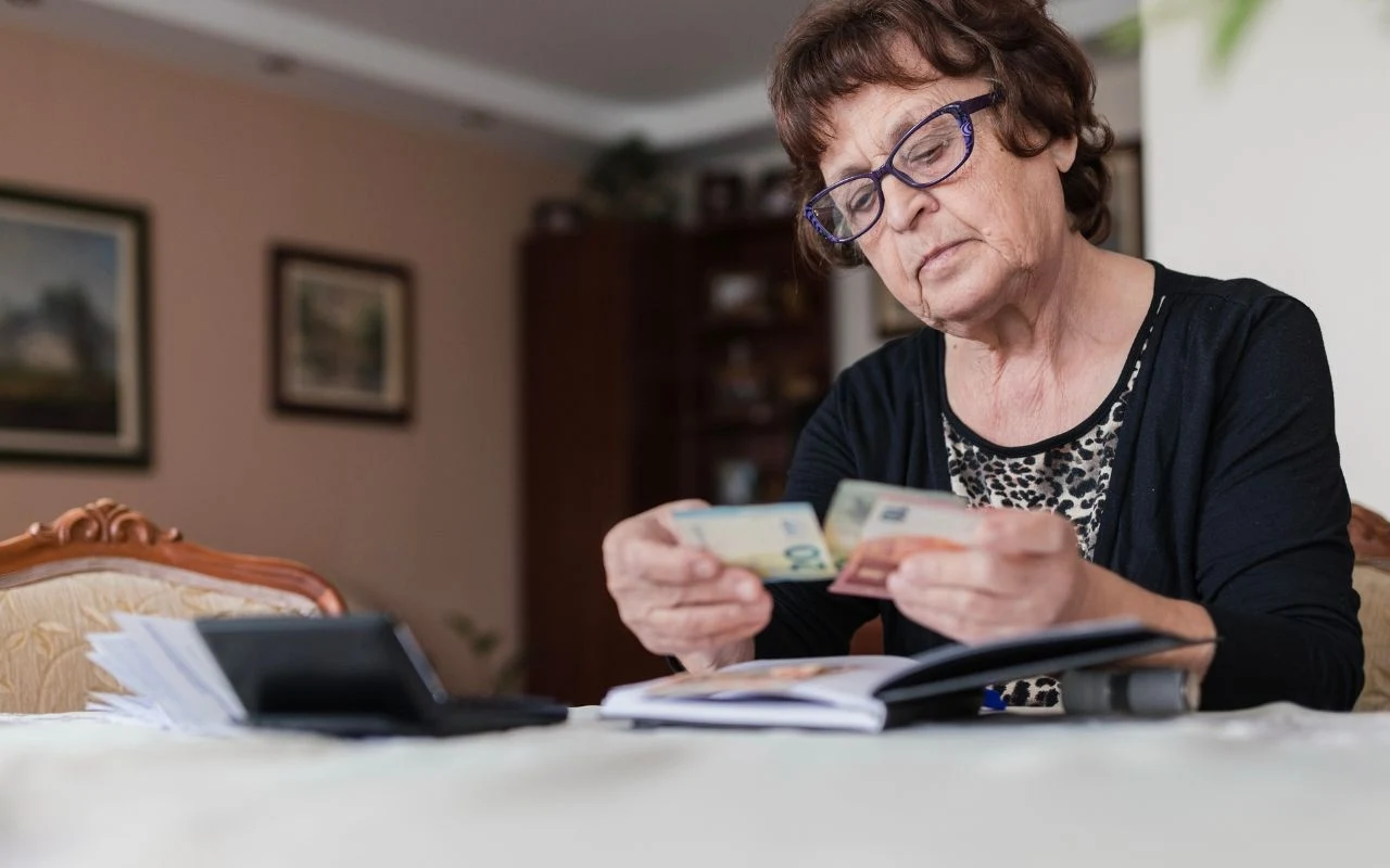 Starsza kobieta w okularach siedzi przy stole, trzyma w rękach banknoty i spogląda na nie uważnie, na stole leżą dokumenty oraz portfel.