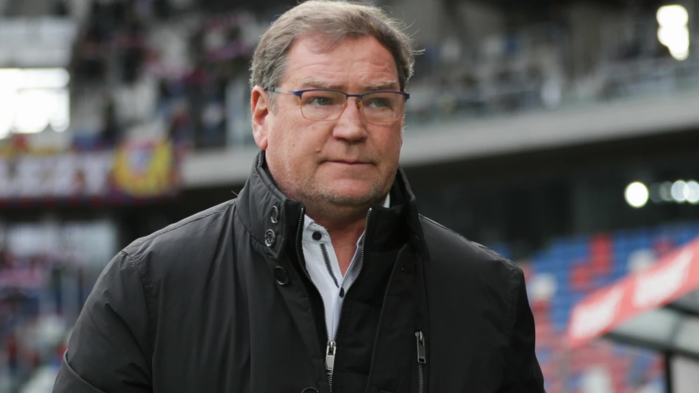 Mężczyzna w okularach w średnim wieku ubrany w ciemną kurtkę i białą koszulę stoi na tle stadionu sportowego.