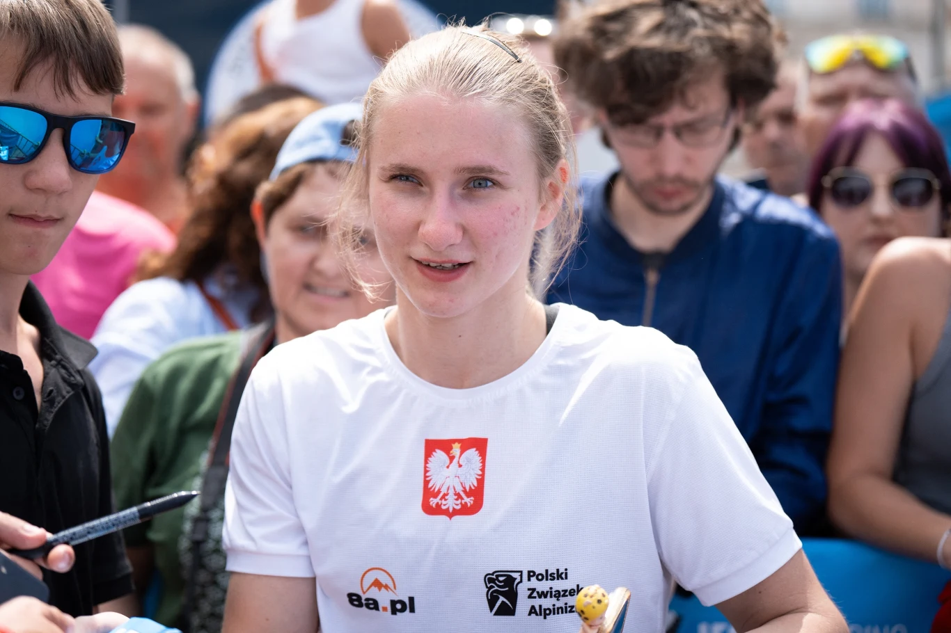 Aleksandra Kałucka Młoda kobieta w białej koszulce z polskim godłem oraz logo Polskiego Związku Alpinizmu, otoczona przez tłum ludzi, z których część ma autografy lub przedmioty do podpisu.