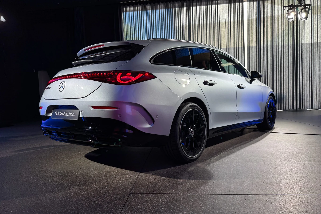Nowy Mercedes CLA debiutuje w bardziej praktycznej odmianie - Shooting Brake Srebrny samochód marki Mercedes, model CLA Shooting Brake, stoi w nowoczesnym wnętrzu z dużymi przeszkleniami i metalicznymi zasłonami. Sportowa sylwetka podkreślona czarnymi felgami oraz włączonymi tylnymi światłami LED.