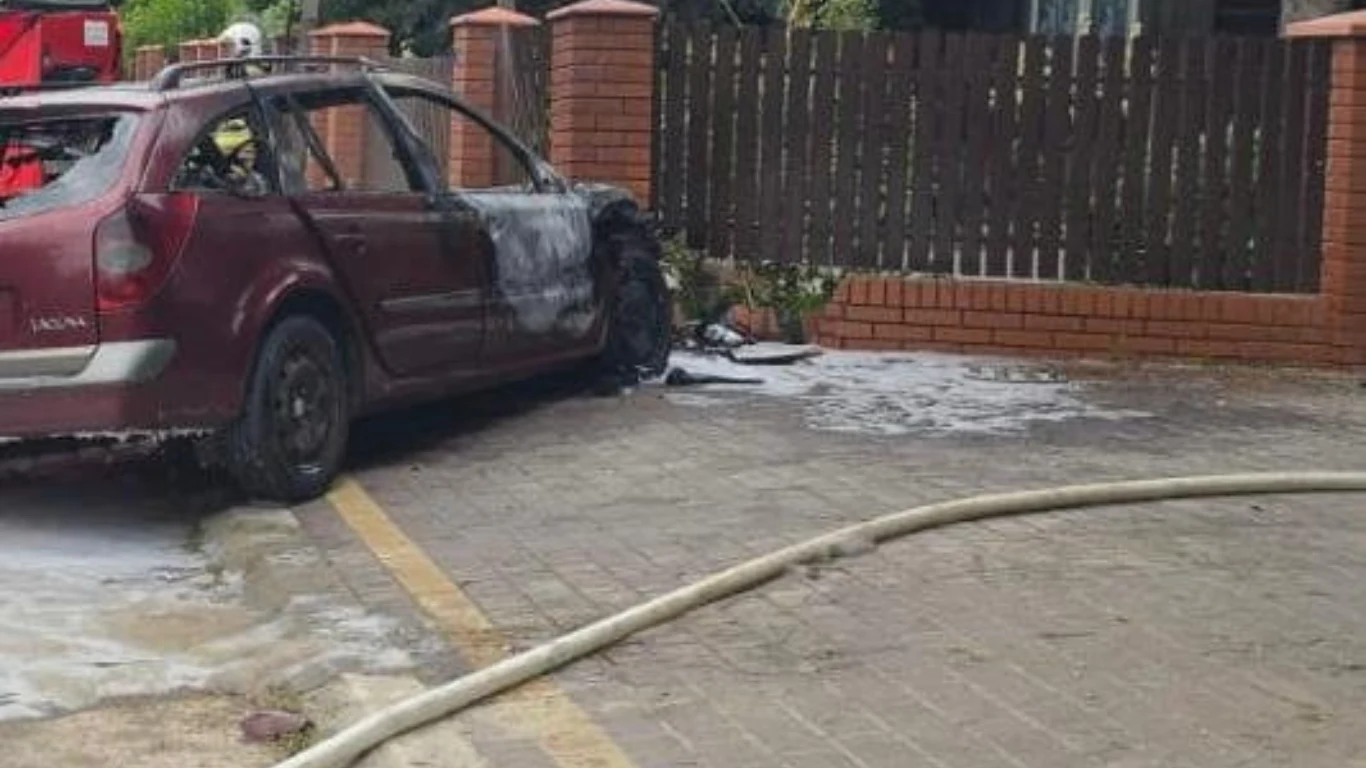 Śmiertelny wypadek w miejscowości Łany Spalony samochód stojący na parkingu przy płocie z czerwonej cegły, widoczne ślady gaszenia pianą, fragment węża strażackiego na ziemi, w tle drewniany płot i roślinność.