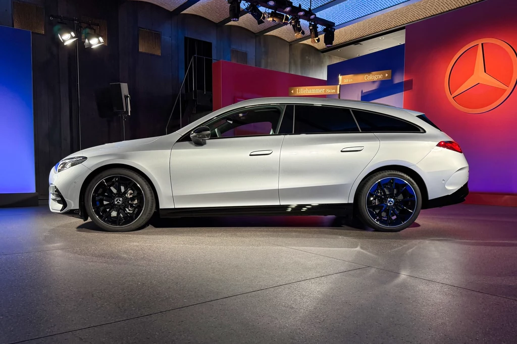 CLA Shooting Brake ma 4723 mm długości, o 35 mm więcej niż model poprzedniej generacji Białe kombi marki Mercedes ustawione w eleganckim, nowoczesnym showroomie, w tle kolorowe panele z dużym logiem Mercedesa.