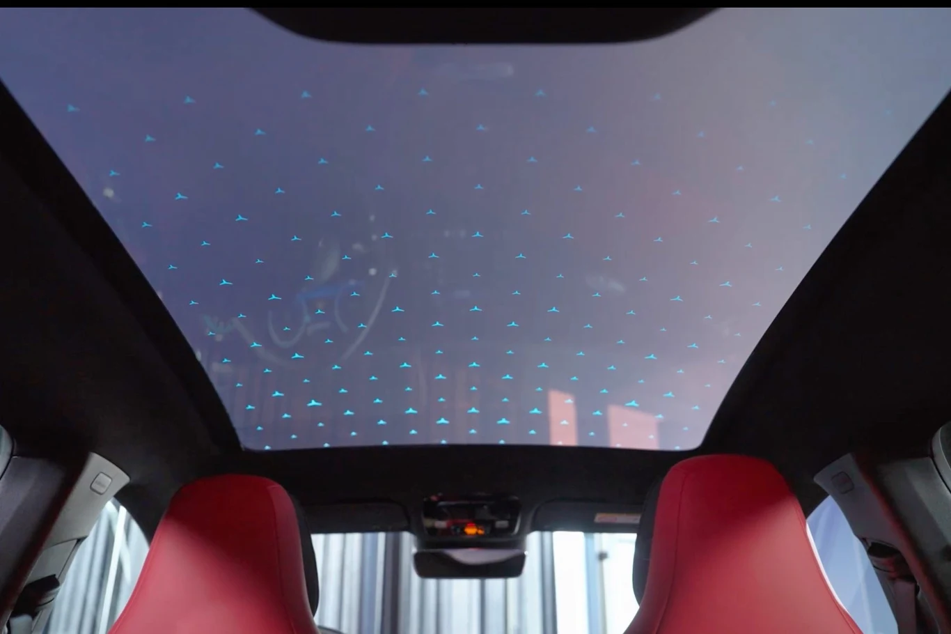 Szyberdach może być przezroczysty lub mleczny. Do tego można podziwiać w nim podświetlone gwiazdy Duży przeszklony dach panoramiczny w samochodzie z podświetleniem LED w kształcie małych niebieskich punktów, dwa przednie fotele wykończone czerwoną tapicerką.