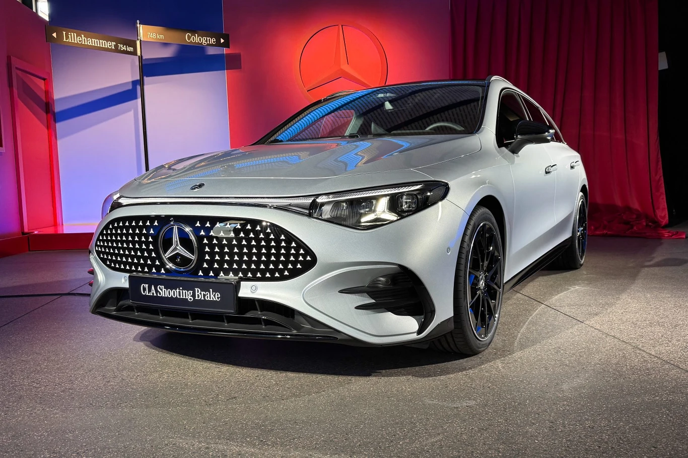 Z przodu auto wygląda jak wersja sedan. Co ważne – w rzeczywistości lepiej niż na zdjęciach Srebrny samochód Mercedes-Benz CLA Shooting Brake stoi na ekspozycji wewnętrznej w otoczeniu czerwonych zasłon oraz tablic kierunkowych, z podświetlonym logotypem marki w tle.