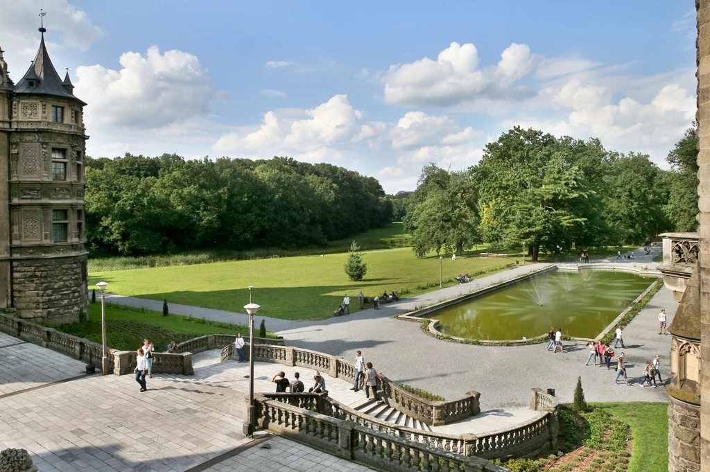 Ogrody zamku w Mosznej Zabytkowy budynek z wieżą, tarasy z ozdobnymi balustradami i staw z fontanną otoczony brukowanymi alejkami, na których spacerują ludzie, w tle rozległy park i drzewa pod błękitnym niebem z chmurami.